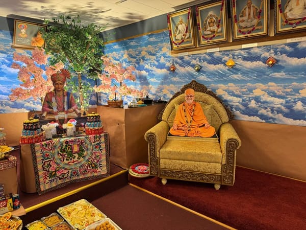 A figure writing the Shikshapatri beside a decorated seat with a life-size saffron-robed cutout, against a painted sky backdrop